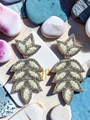 A pair of large, silver earrings with white leaf-like designs and sparkling rhinestones, placed on a surface with various other objects such as seashells and rocks.