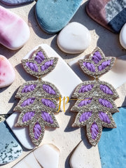A pair of earrings with purple and silver leaf-shaped designs, placed on a surface with various other objects such as rocks and seashells.