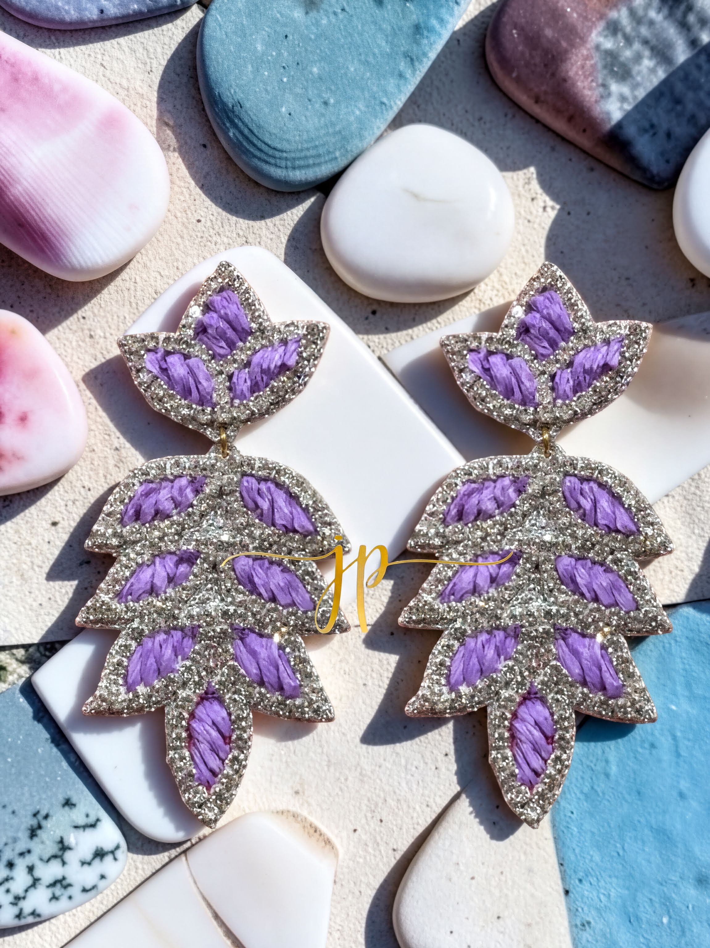A pair of earrings with purple and silver leaf-shaped designs, placed on a surface with various other objects such as rocks and seashells.